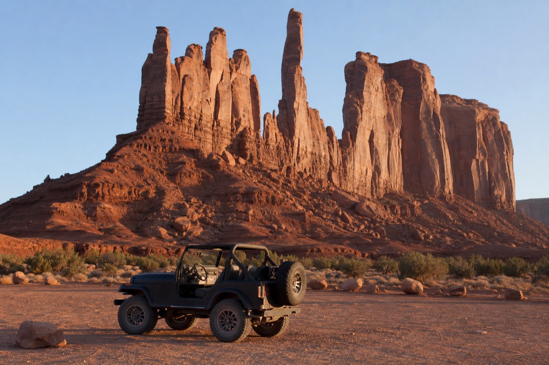 An open-air jeep parked on the sandy valley floor beneath towering sandstone spires in golden-hour light