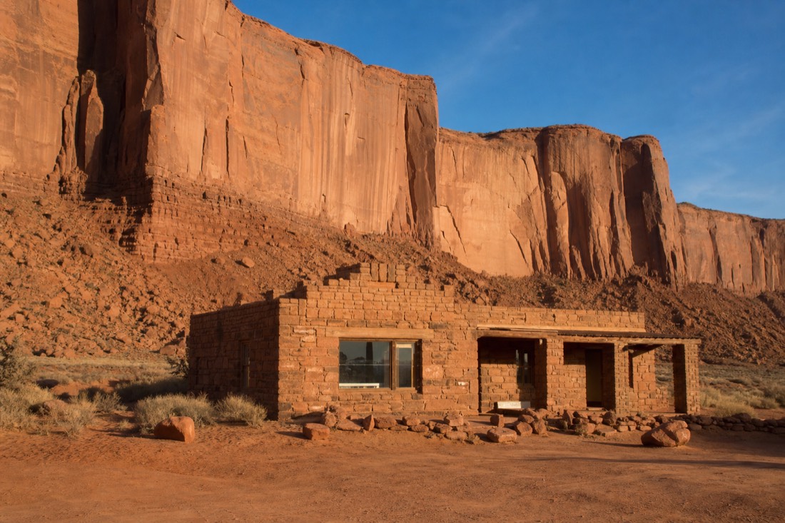 A weathered stone trading post building below towering red sandstone cliffs at the edge of Monument Valley in warm afternoon light