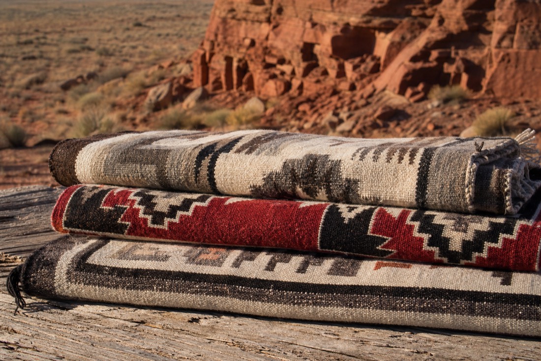 Three folded handwoven Navajo rugs in Two Grey Hills, Ganado red, and bordered patterns stacked on weathered wood