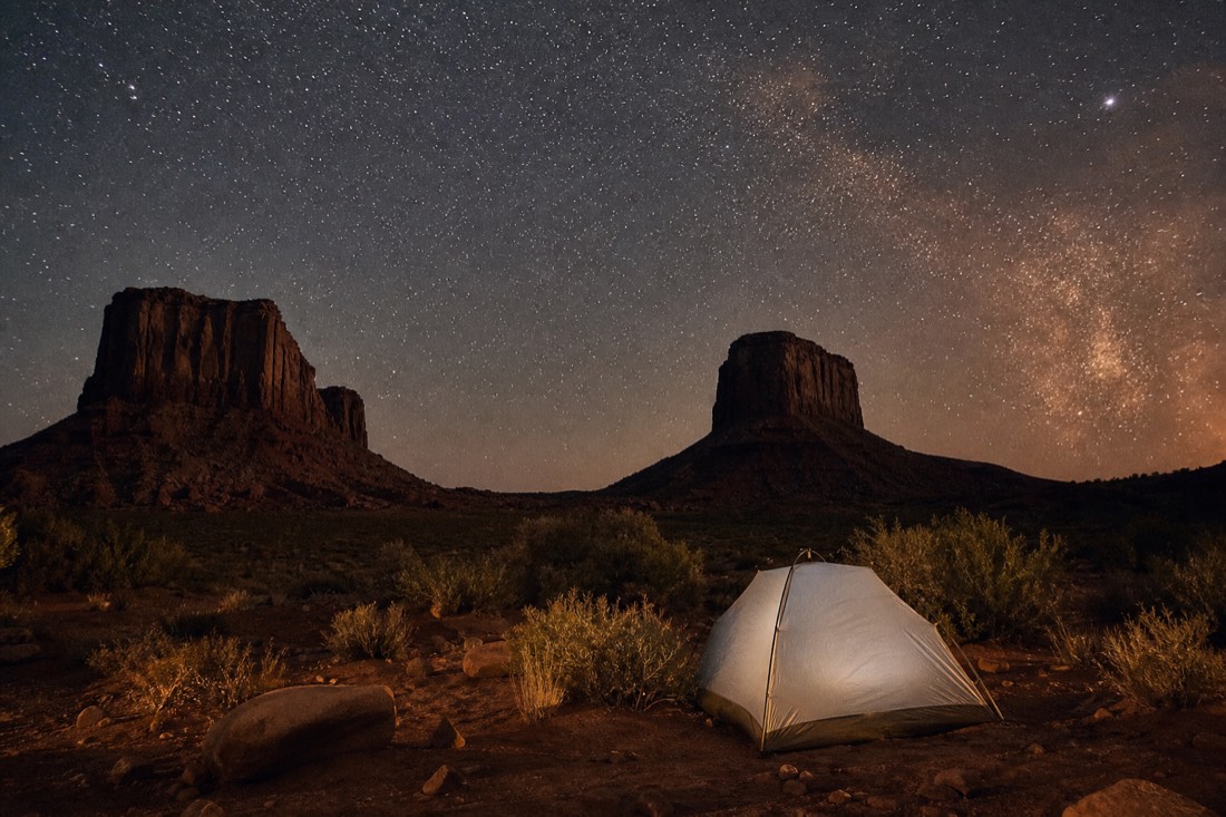A quiet tent campsite on the valley floor beneath a sweeping field of stars, buttes standing black against the night