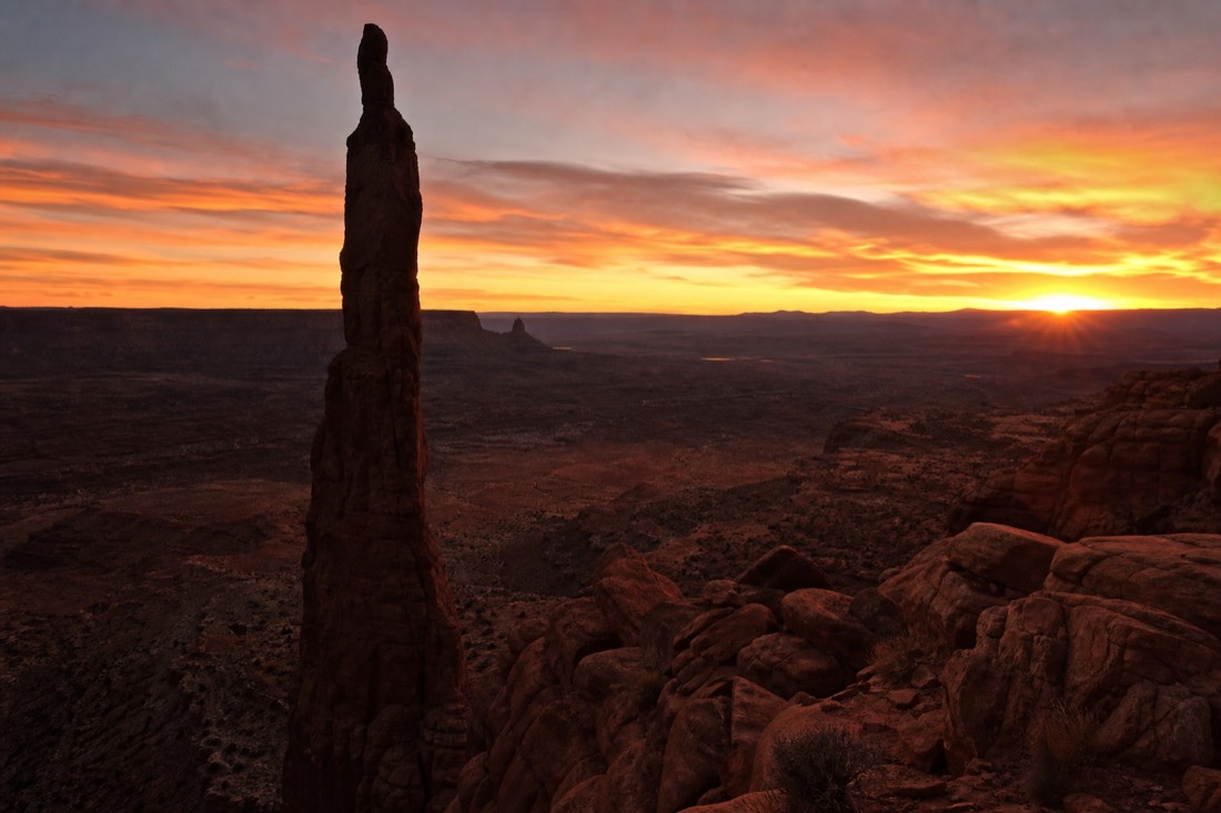 The thin Totem Pole spire silhouetted against a glowing orange and pink sunrise sky over the dark valley floor