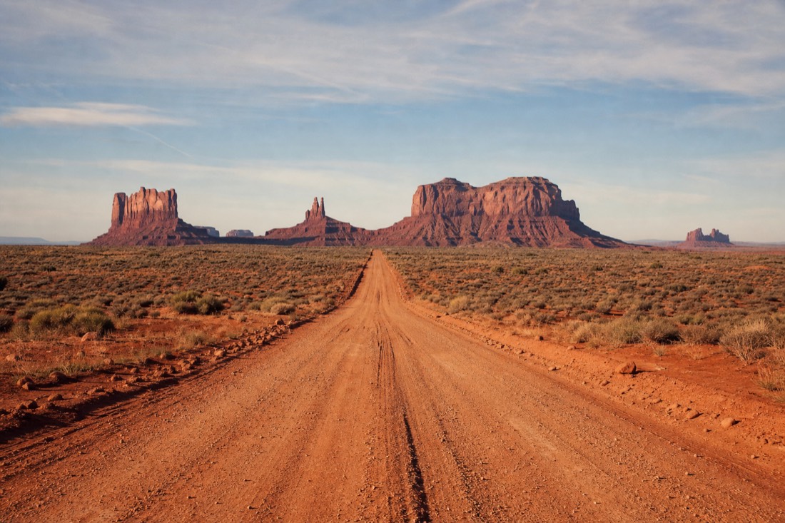 A marked red dirt road running straight across the Monument Valley floor toward distant sandstone buttes