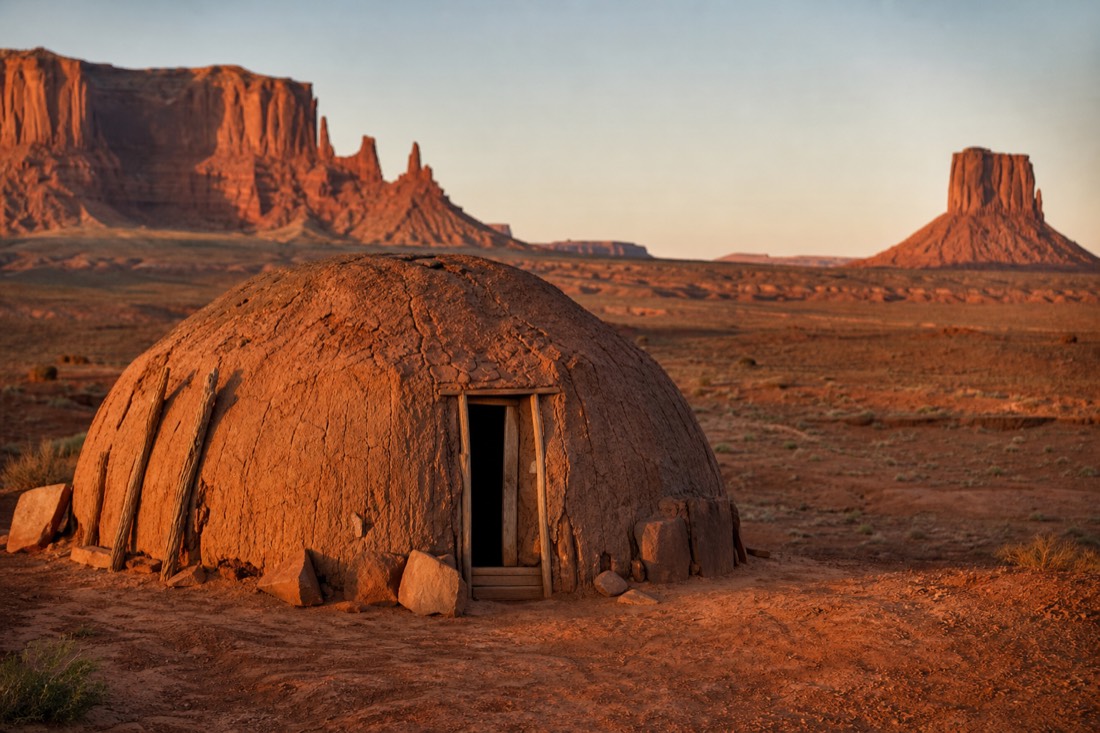 A round timber-and-earth Navajo hogan on the red desert floor beneath Monument Valley buttes at golden hour
