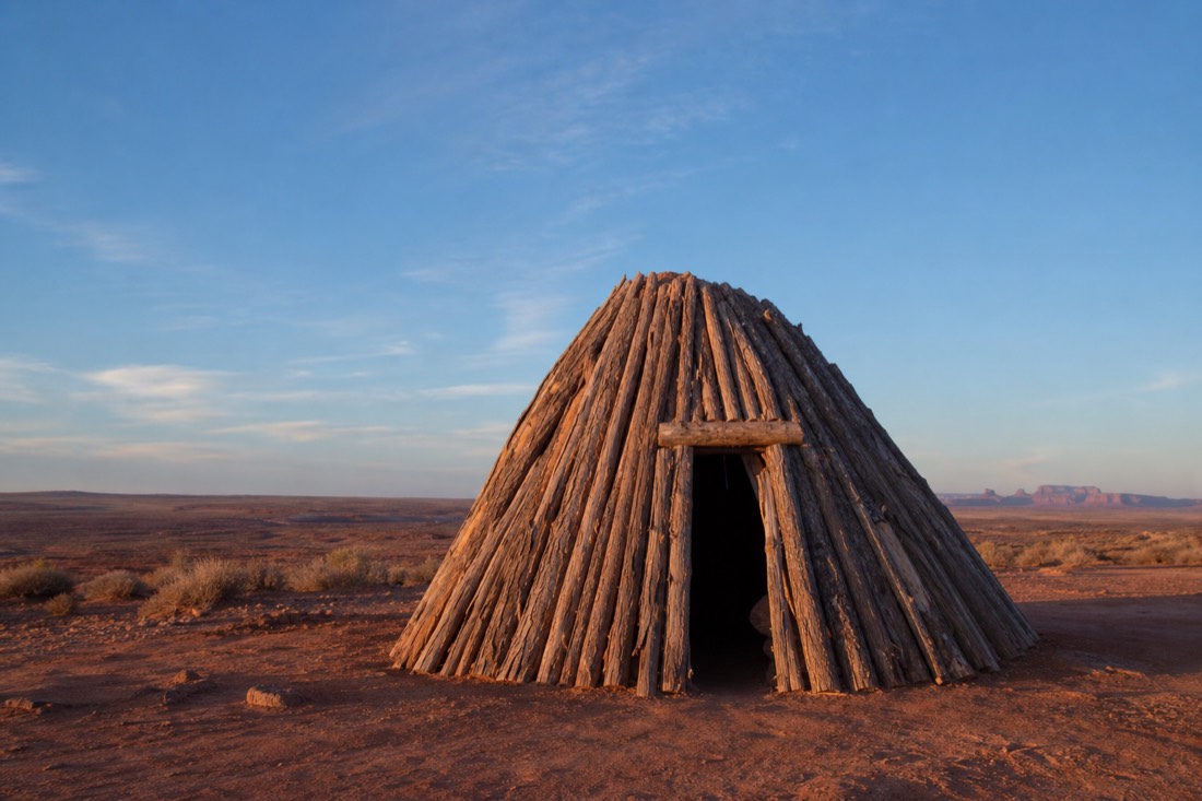 A cone-shaped forked-stick hogan of stacked logs standing alone on open red sand under a wide blue sky