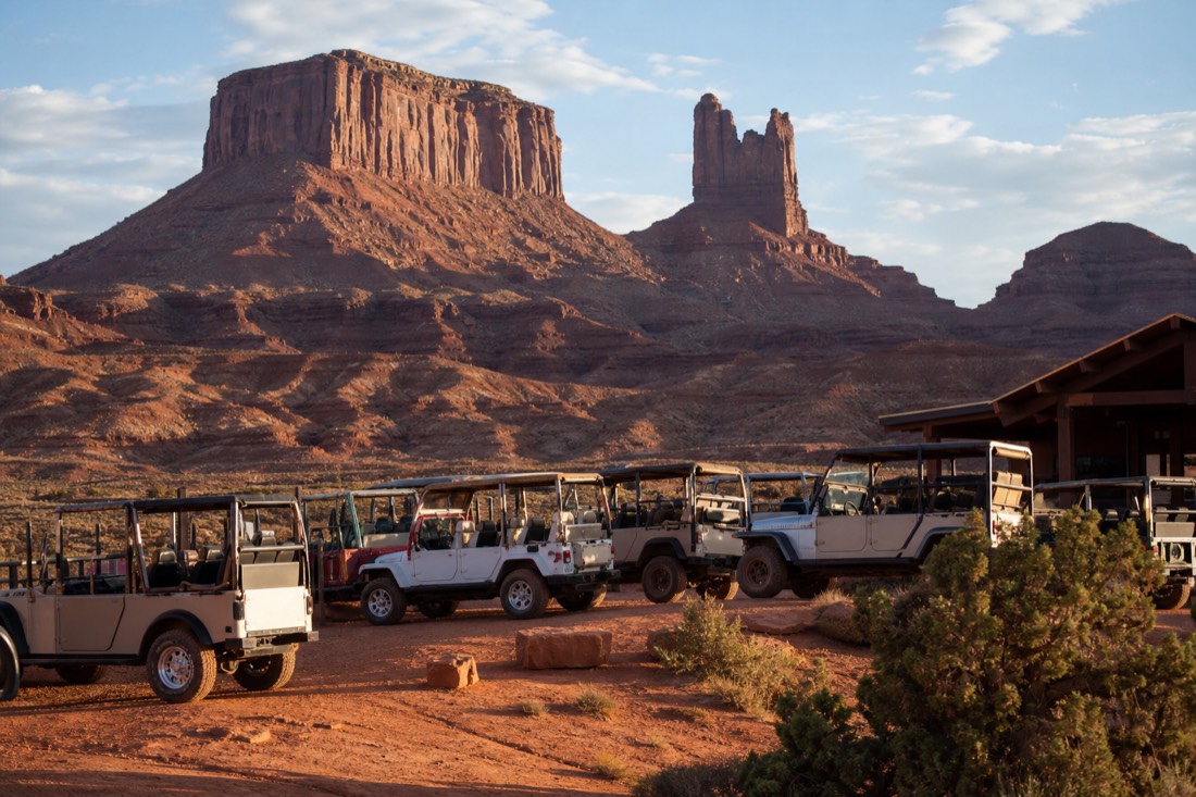 Several open-air tour jeeps lined up on red dirt near the visitor center rim with the Mitten Buttes behind