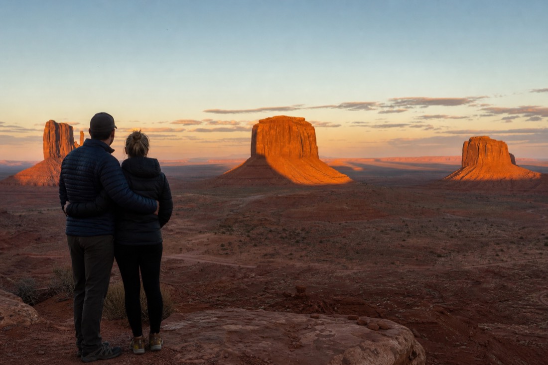 A couple silhouetted at an overlook watching golden sunset light spread across the Mitten Buttes