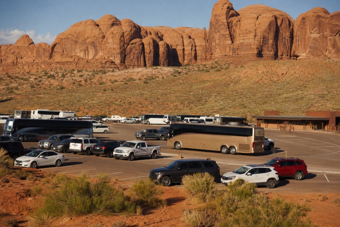 A full parking area of tour vehicles and rental cars beside the visitor center on a busy clear summer morning