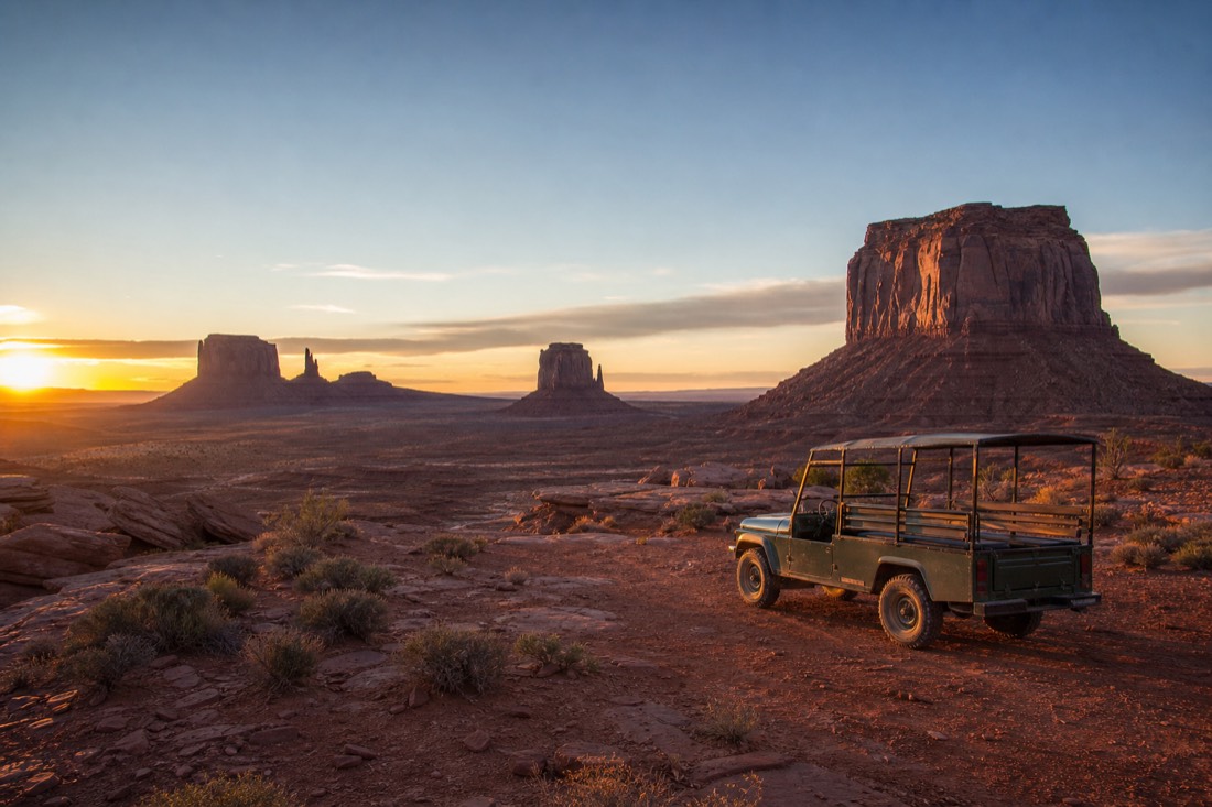 First light breaking over the buttes at dawn with an open-air Navajo jeep waiting on the red sand