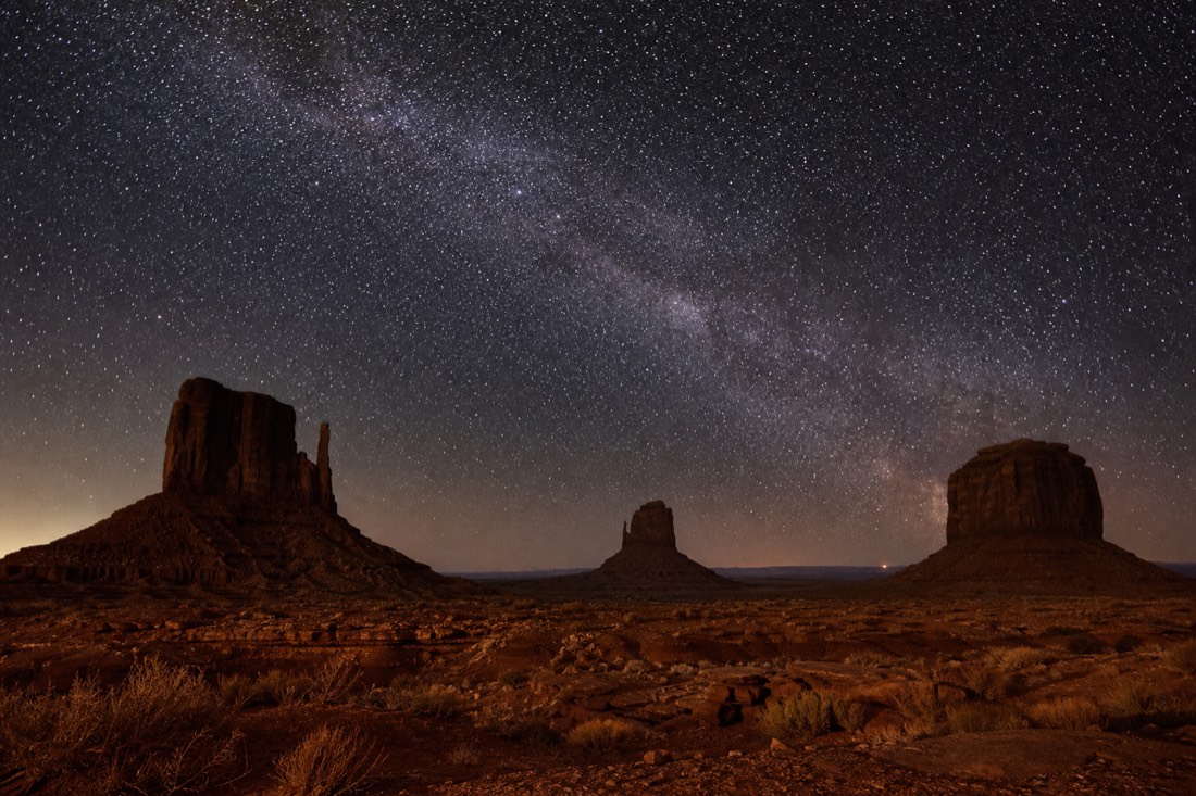 A dense star field and faint Milky Way arching over the dark silhouettes of the Mitten buttes on a clear moonless night