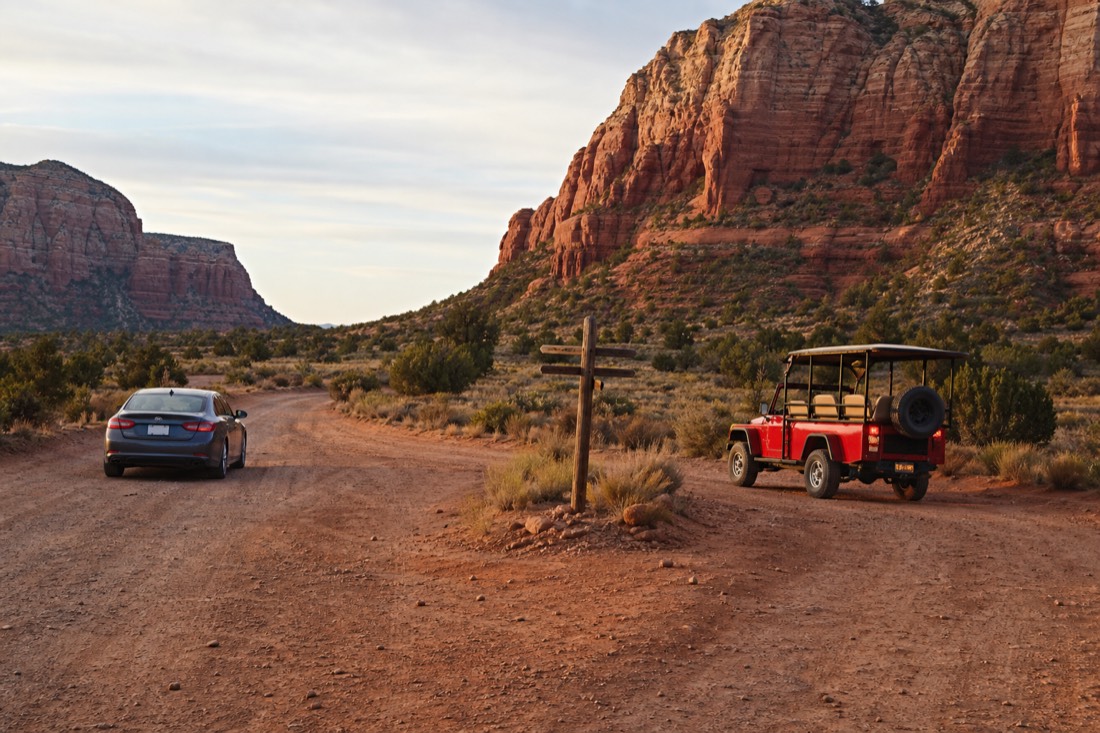 Two vehicles at a fork on the dirt loop, one private car and one open-air tour jeep, red buttes and a weathered signpost between them