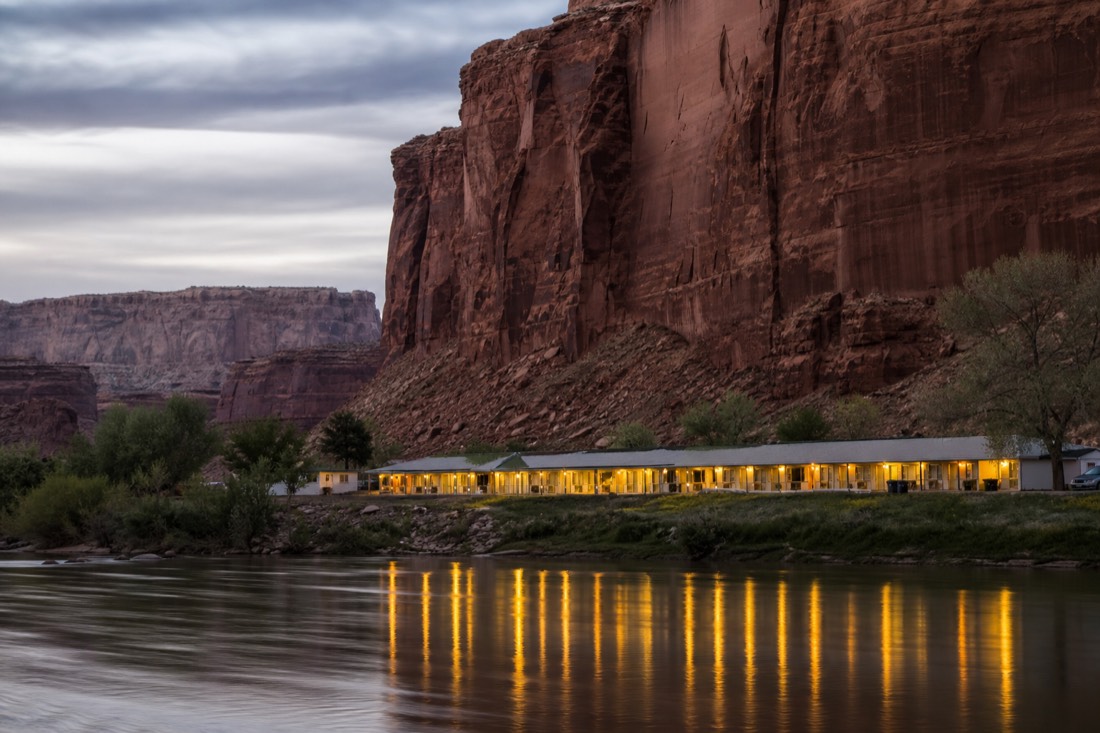 A quiet riverside motel at dusk beneath towering red cliffs, warm window lights reflected in the slow San Juan River