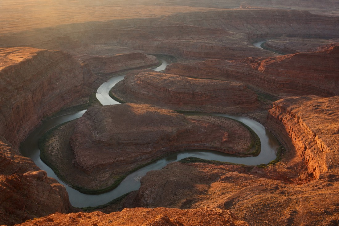 An aerial-style view of a river carved into tight looping horseshoe meanders deep in a bare desert canyon at golden hour