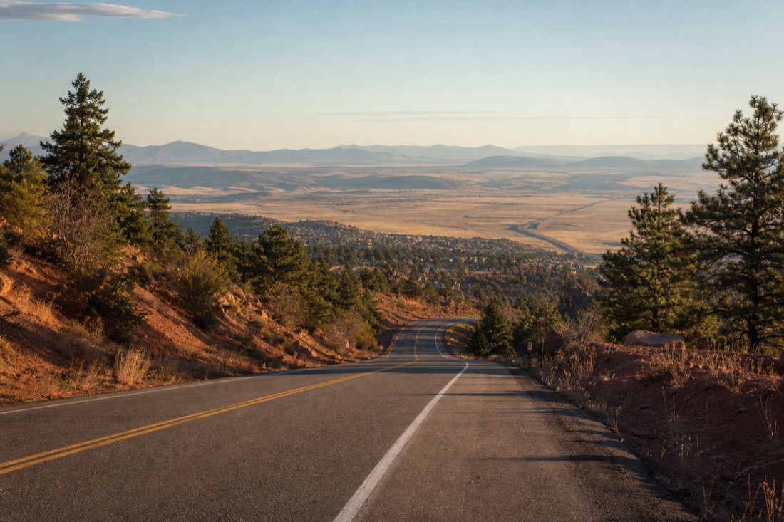 A highway leaving a pine-forested mountain town and descending toward open high-desert grassland in morning light