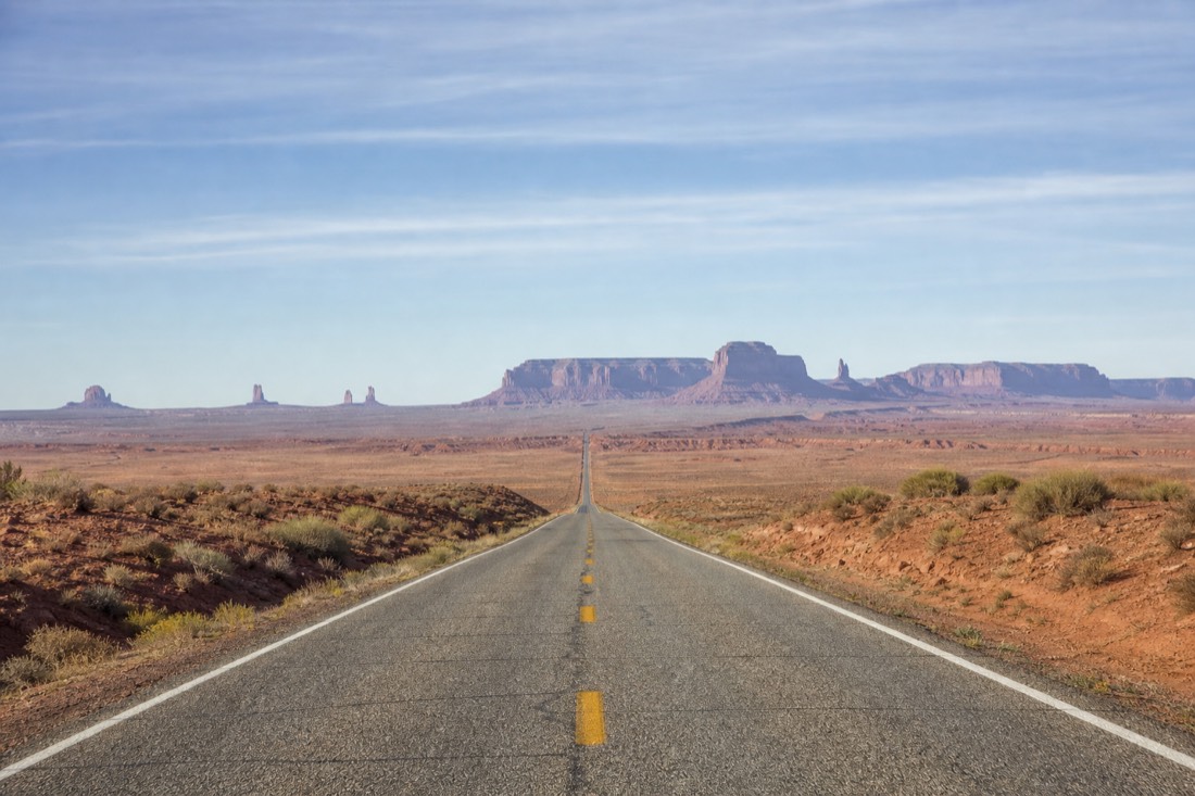 An empty paved highway running south across flat red desert toward distant blue mesas under a wide morning sky