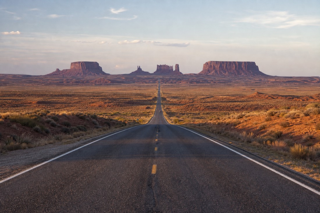 US-163 running dead straight toward Monument Valley's lined-up buttes on the horizon from the classic northern approach