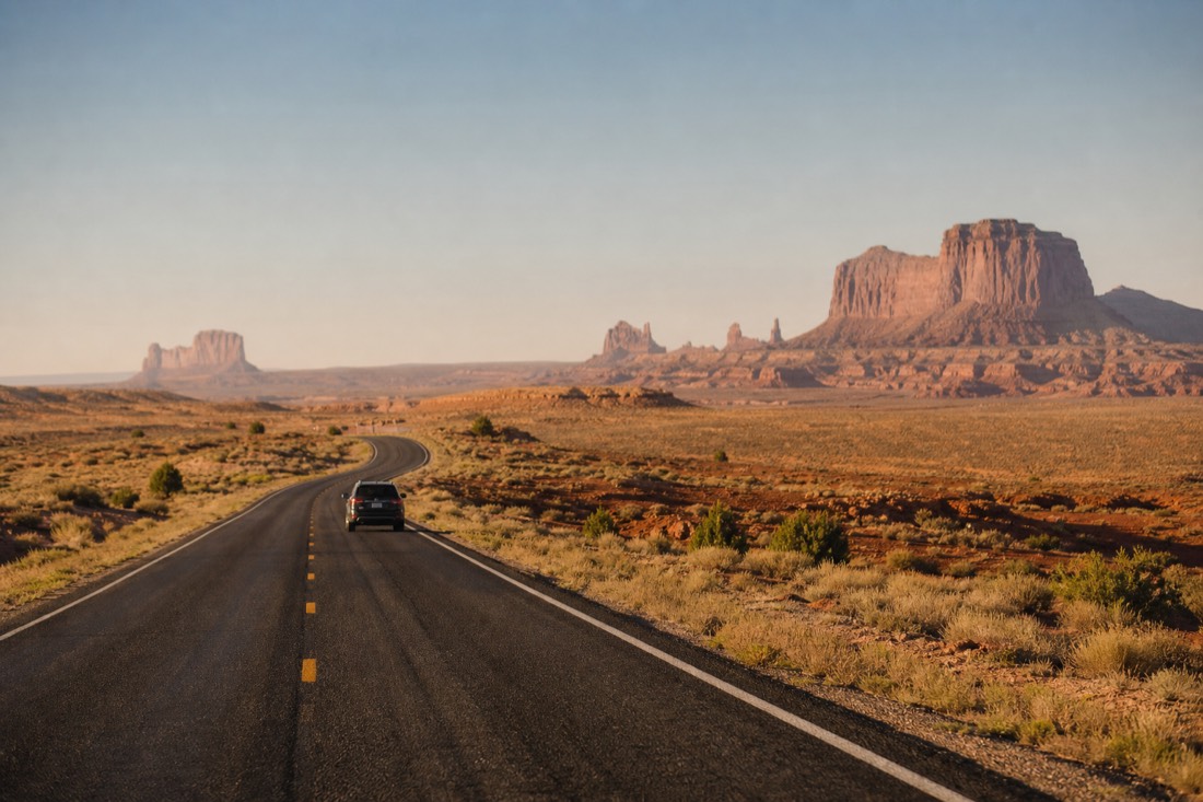 A lone car on a quiet two-lane desert road with sparse scrub and far-off red rock formations shimmering in heat haze