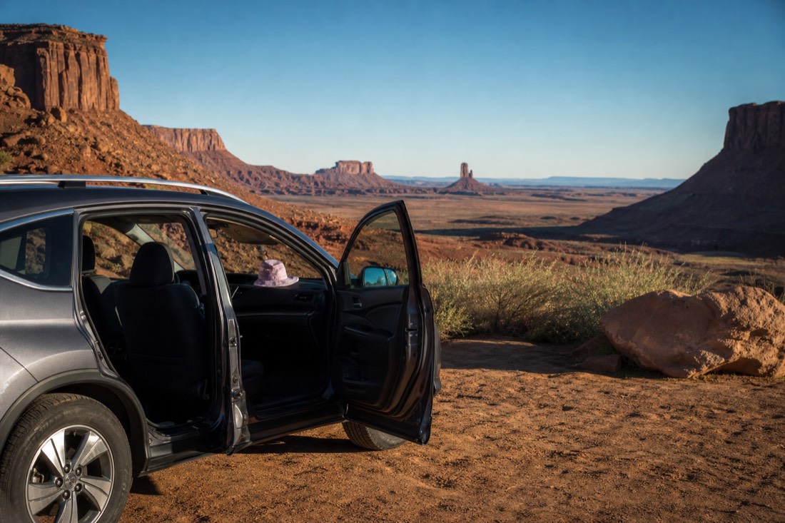 A parked crossover SUV with its doors open at a valley overlook, a child's sun hat resting on the dashboard and buttes beyond