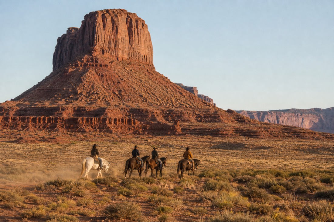 Horseback riders crossing the open valley floor beneath Merrick Butte in warm late-afternoon light