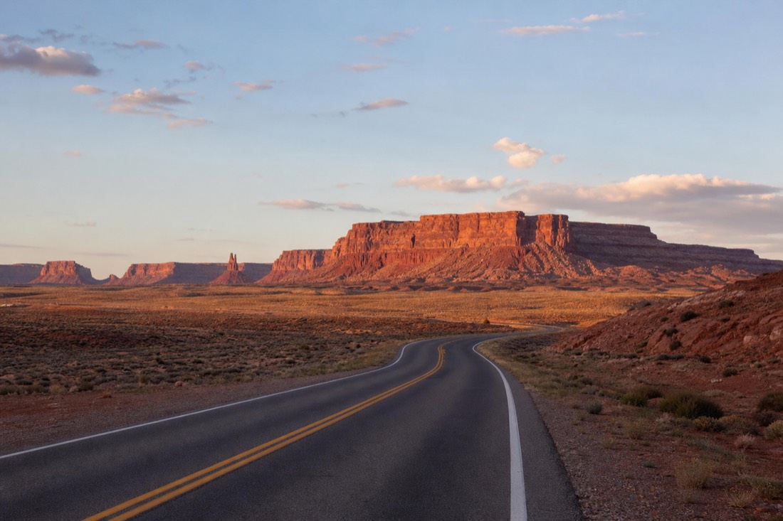 An empty desert highway curving toward layered red mesas under a wide Southwest sky at golden hour