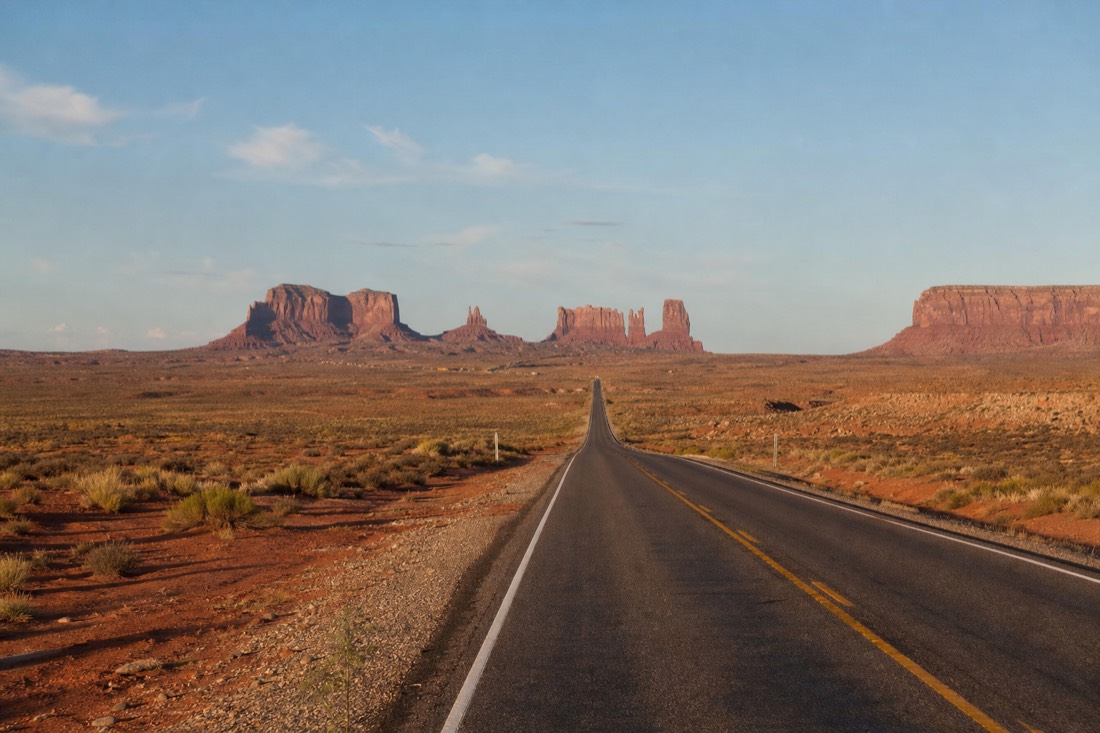 A straight red-dirt-lined highway running toward tall sandstone towers in the clear midday distance