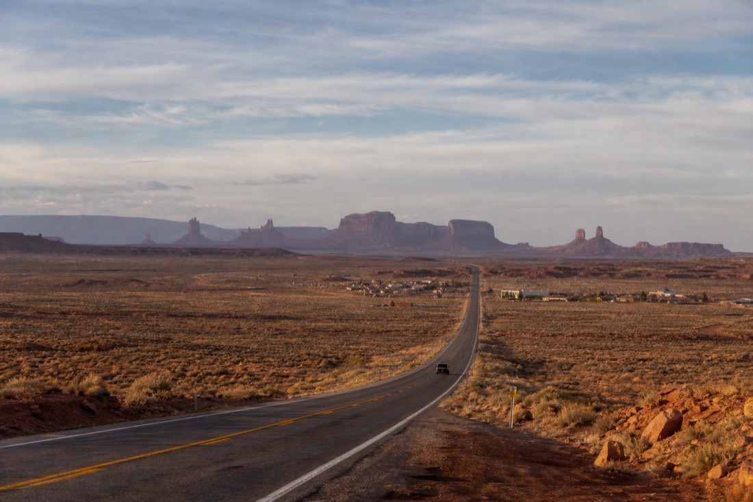 US-163 highway crossing open high desert toward a small roadside motel town, Monument Valley buttes on the far horizon