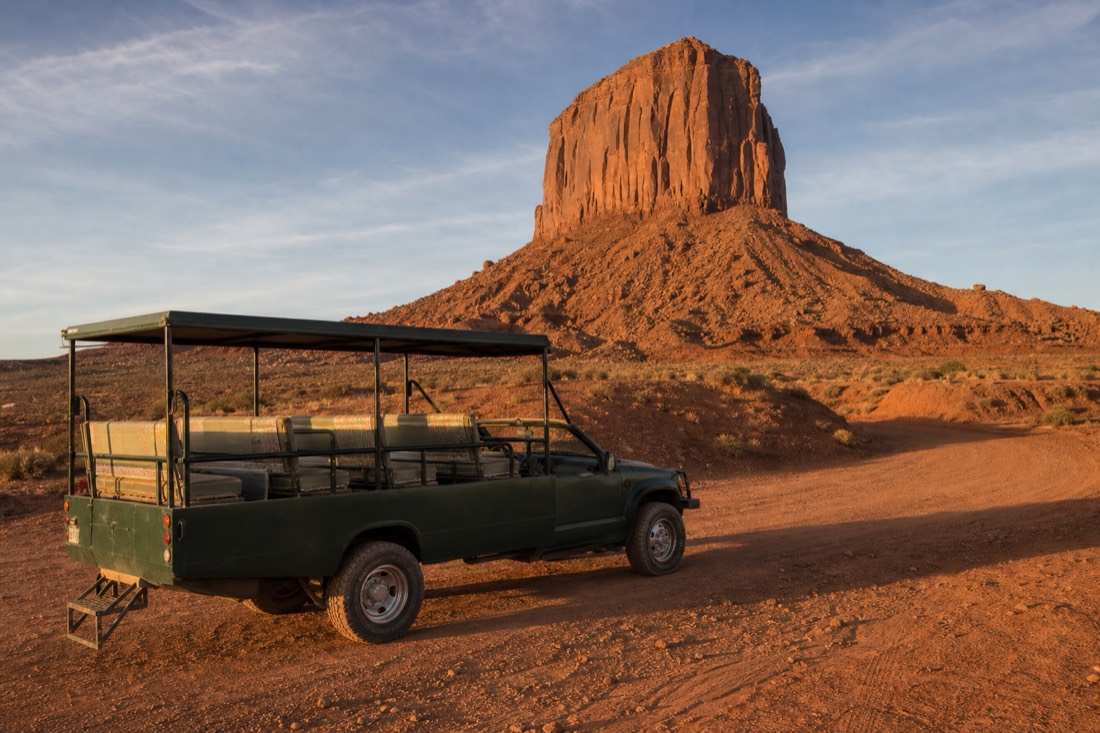 Open-air Navajo tour jeep on the red dirt loop road at golden hour with Merrick Butte rising behind