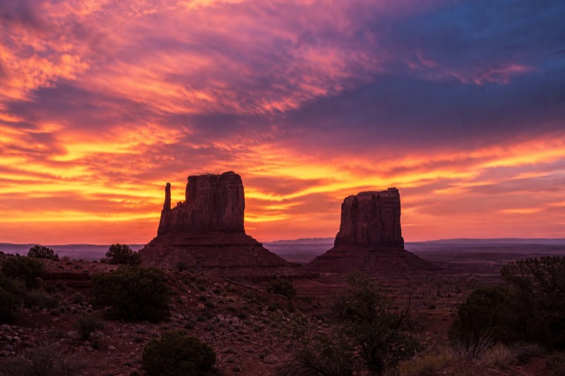 The Mittens silhouetted against a burning orange and violet sky at sunset in Monument Valley
